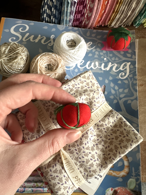 A hand holding a mini tomato pin cushion. Under is a sewing book, spools of cotton thread and stacks of a fabric.