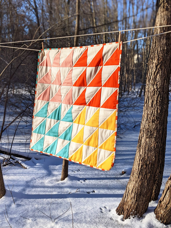 A quilt with teal, yellow, peach and cream triangle design hangs on a rope in the woods.