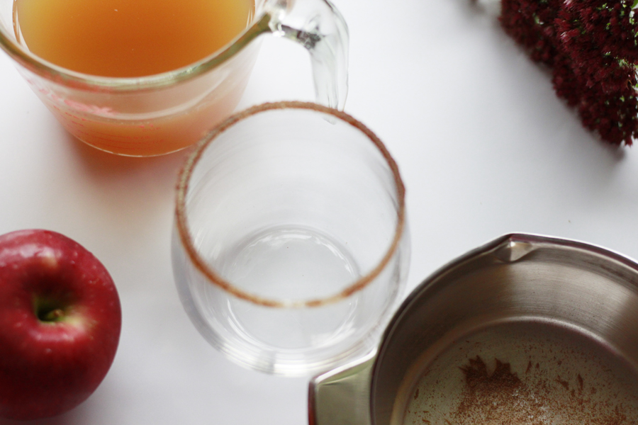 The picture taken from above vantage point. Apples scatterd on the table with measuring cup filled with apple cider. A glass with cinnamon sugar on the rim, and a cup up of cinnamon sugar 