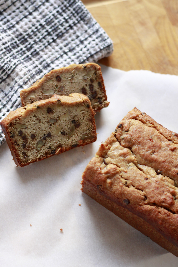 two slices of peanut butter chocolate chip banana bread and a loaf sitting on parchment paper, a black and white dishcloth and a wooden cutting board.