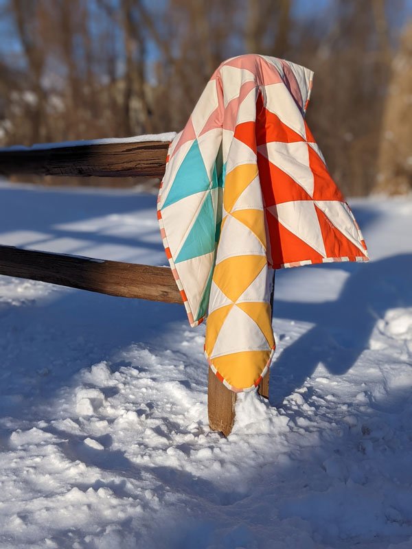 A rustic wooden fence with a quilt with bright colors hanging on it. It's snowy and there are woods in the background 