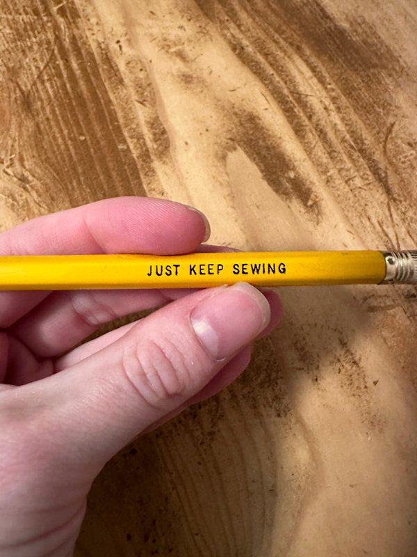 hand holding a yellow, wooden pencil that says "just keep sewing"