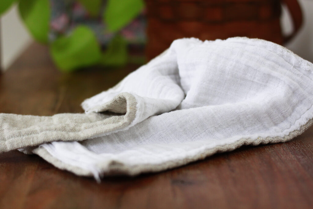 A biege homemade napkin with one side warm grey and highly textured, drop cloth  the other side a white guaze fabric. 