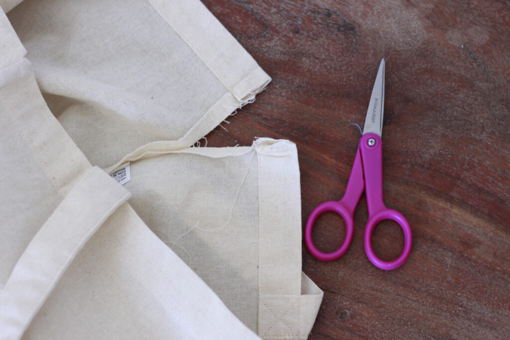 A seam open on a plain canvas bag, small, sharp pink scissors are next to it. 