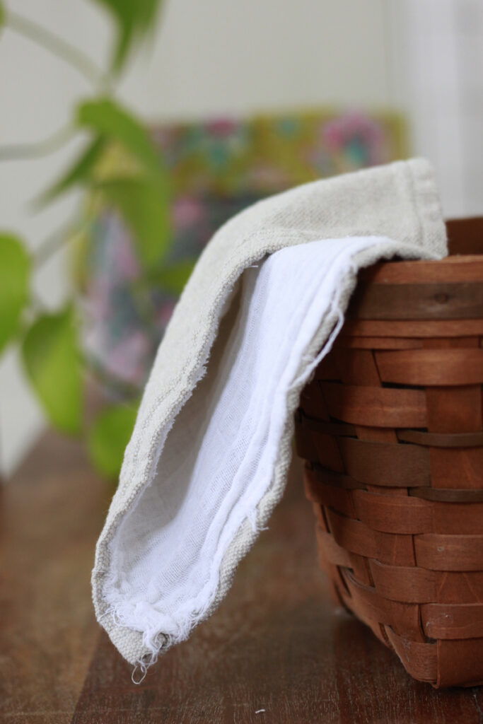 In a basket, a biege homemade napkin with one side warm grey and highly textured, the other side a white guaze fabric hangs off the side. In the background there is a plant and colorful blurry objects