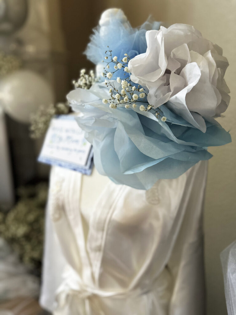 A very large blue paper flower and small white flower, baby's breath and tuft of blue tulle are arranged on a manikin. The mankin has a silk white robe with lace. In the background there is an illegable card, blurry balloons and babys breath flowers.