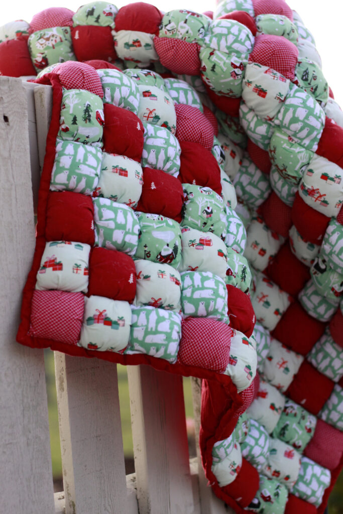 A puffy blanket with Christmas color palette, prints have polar bears and christmas presents, the blanket hangs on a rustic fence