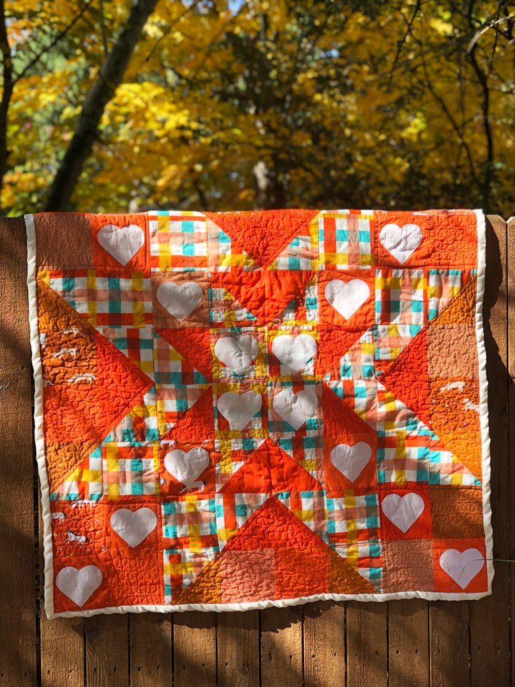 A bright orange quilt hands from a fence, the light is dappled and the quilt has heart applique and horse prints.