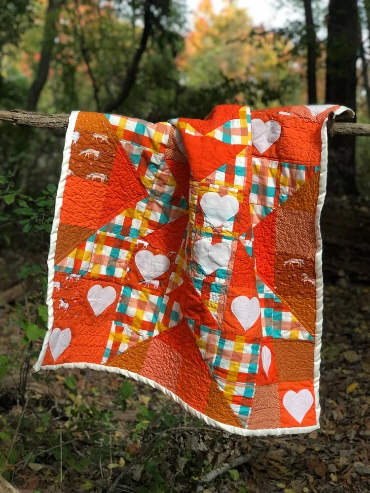 An orange quilt is shown on a stick, it has a silk edging and heart applique. There are variation in the quilt that show the stitches from far away.
