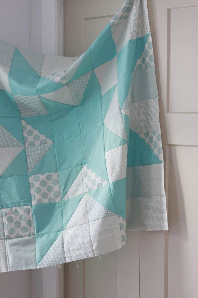 A sawtooth star quilt top in cream and teal hang on in a door way with white walls. 