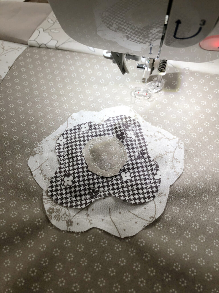 Shown under a sewing machine's footer, three layers of a cream and white floral fabrics create a quilted flower with white thread. 