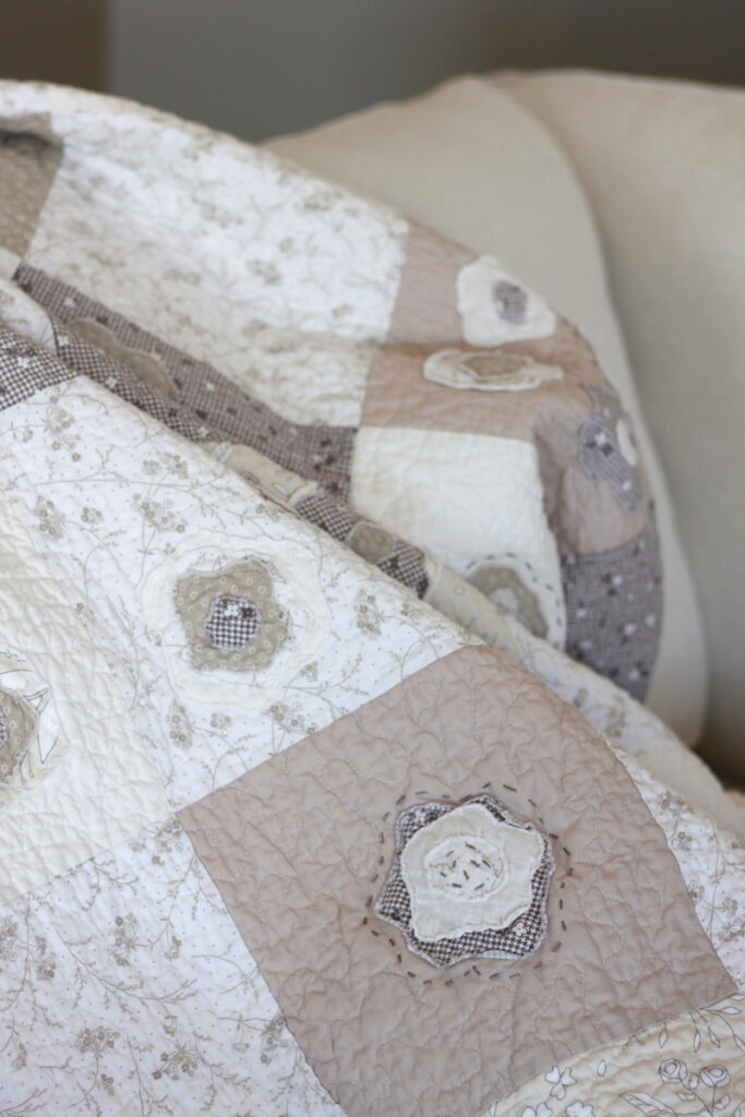 A quilt draped over a couch showing detail stitching around a rose. The quilt blocks are all in cream, whites and light browns.